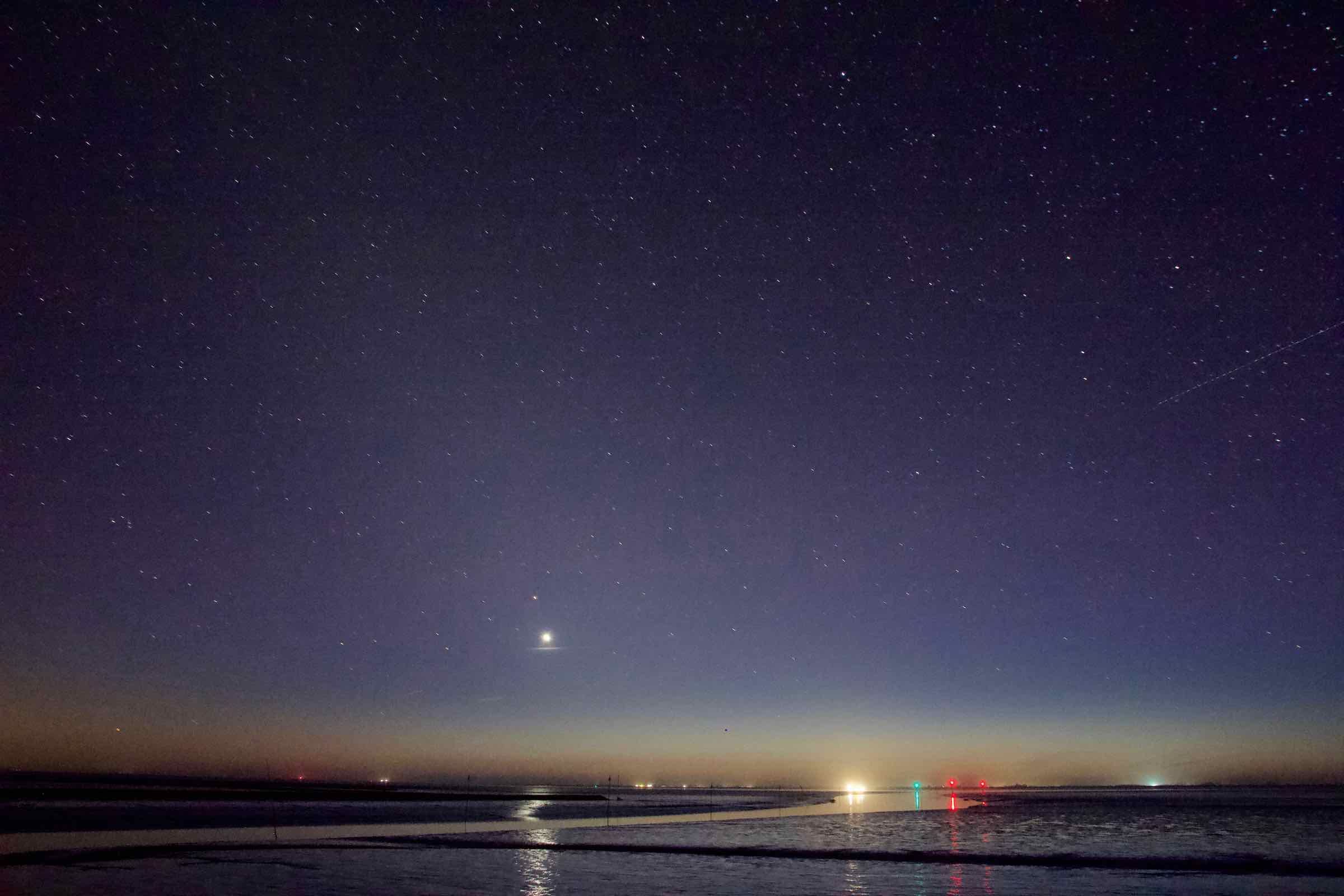 Nachts erscheinen am Meer vor dem Dockkoog viele Seezeichen und darüber ein riesiger Sternenhimmel. 