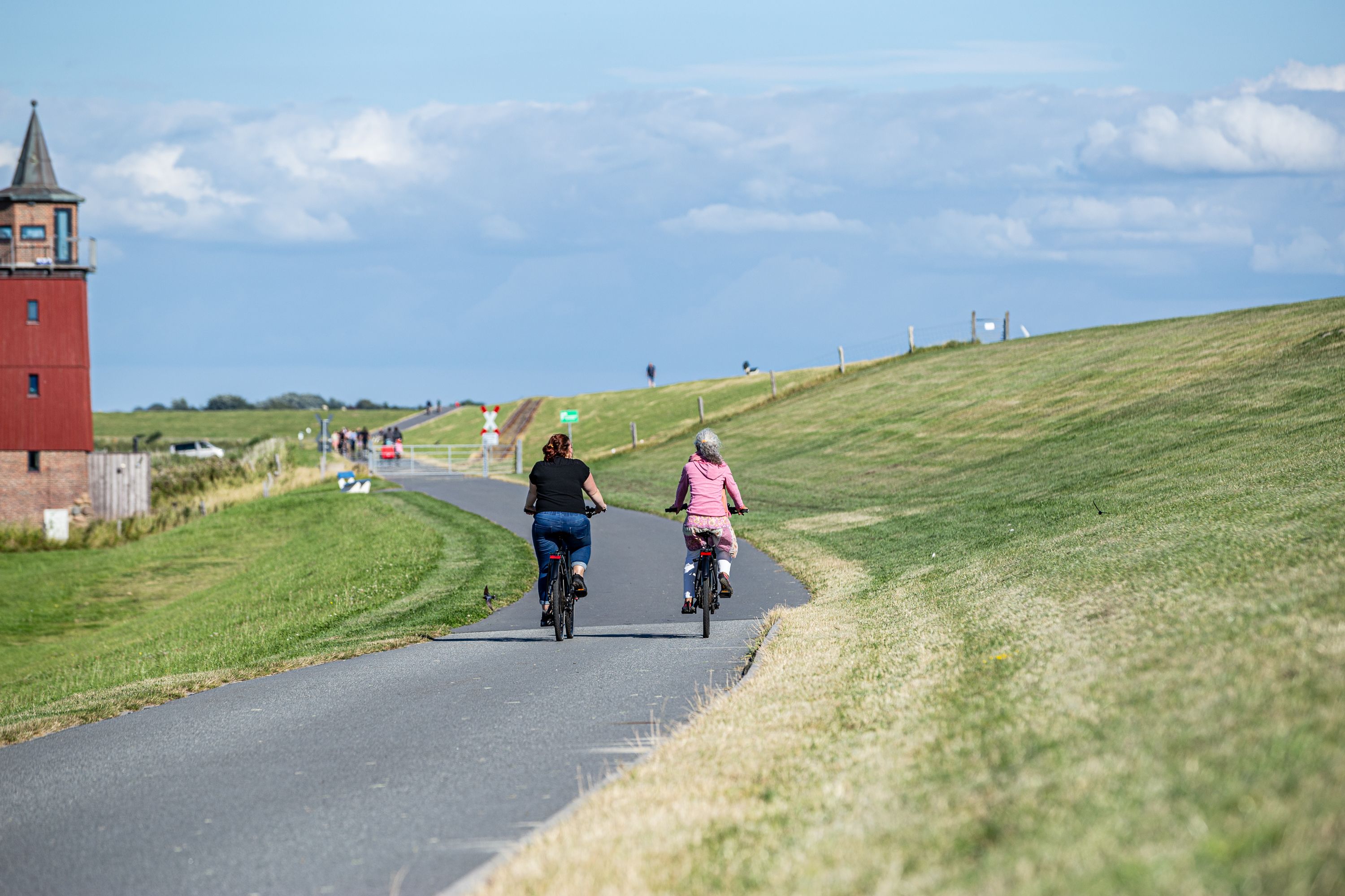 Zwei Personen fahren am Deich Fahrrad, im Hintergrund steht der Dagebüller Leuchtturm